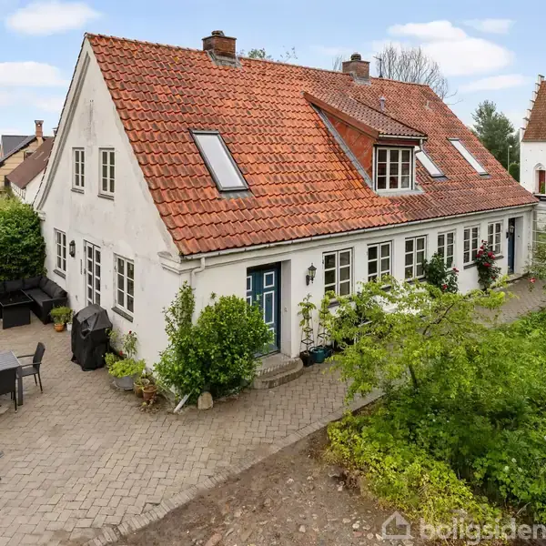 Hvidt hus med rødt tegltag har flere vinduer og en blå dør. Det står i en have med fliser og havemøbler. I baggrunden ses en bygning med kirkelignende træk.