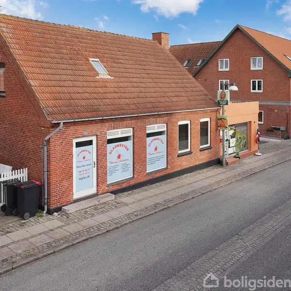 En rød murstensbygning med trekantet tag har butiksvinduer med teksten: "Afrodite Klip & Style, Vi har åbent alle dage fra kl. 9:00 til kl. 18:00." Bygningen står på en stille gade.