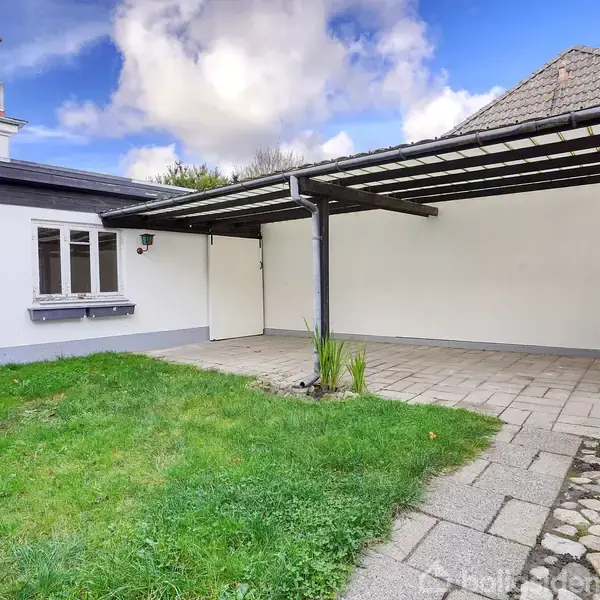 Carport med fladt tag og åbne søjler ved siden af et hvidt hus. Området inkluderer en belagt terrasse, grønt græs og dekorationer mod væggen.
