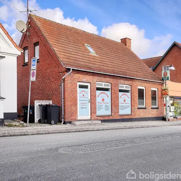 En rød murstensbygning med tegltag, der fungerer som en virksomhed. Vinduer og indgang er udsmykket med tekst: "Pizzeria Italiano Borsholm," og adressen "Nygade 12A" ses blandt anden tekst. Bygningen ligger ved siden af en vej i et byområde.