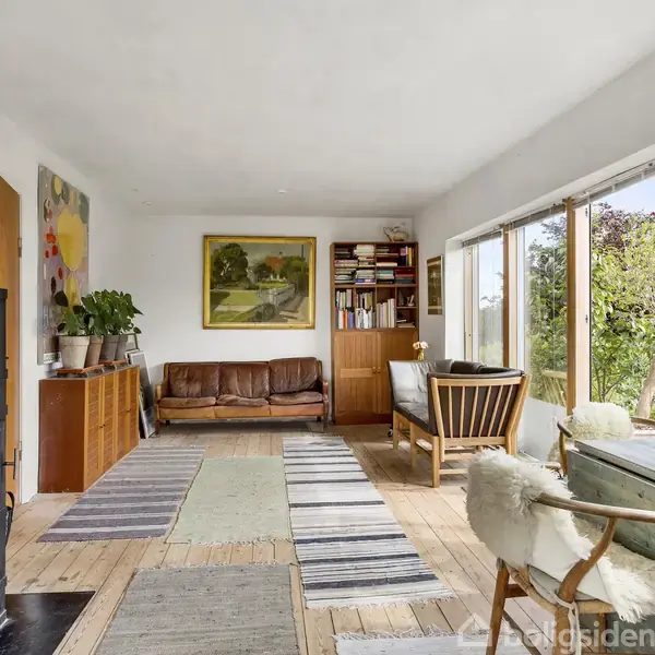 En stue med brændeovn til venstre, brændende. Træmøbler og sofaer står langs vinduerne med udsigt til en grøn have. Hylder med bøger og planter dekorerer rummet.