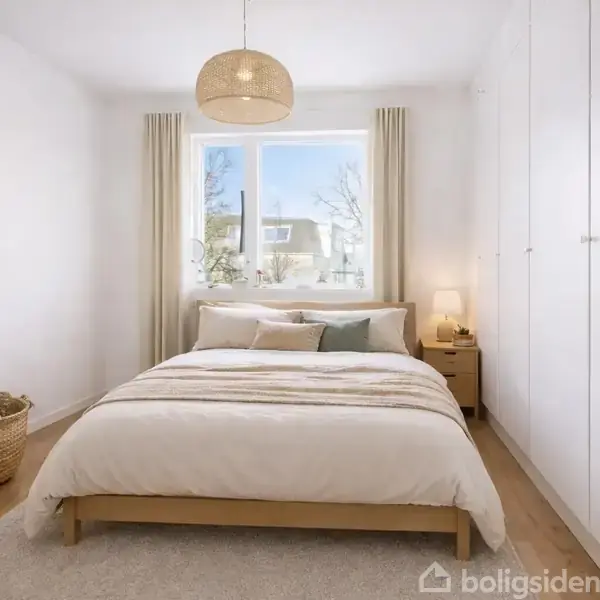 Seng står i midten af et lyst soveværelse med hvide vægge. Ved siden af er et natbord med lampe. Et stort vindue med gardiner lader lys komme ind.