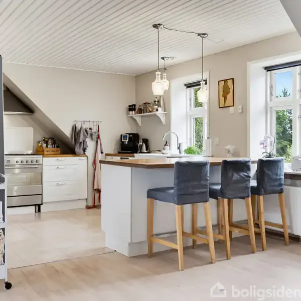 Et moderne køkken med en kogeø, tre barstole og stilfuld belysning. Køkkenet har lyse træmøbler og hvidevarer, med et vindue, der giver naturligt lys og udsigt til haven.