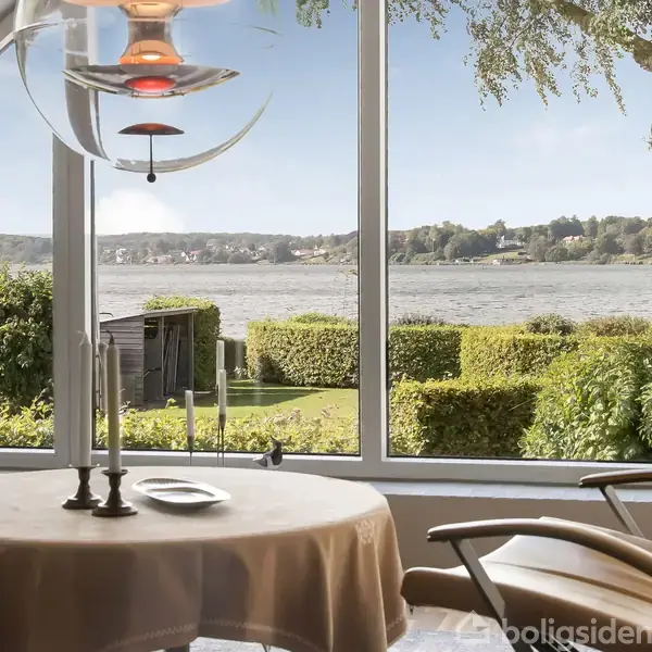 En stol står ved et rundt bord med stearinlys, placeret foran en stor bueformet vindue med udsigt over en sø og grønne buske, under en moderne loftlampe.