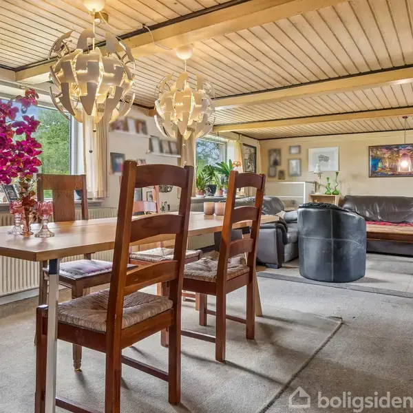 Spisestue med et træbord og stole, omkranset af hvide lamper hængende fra loftet. Til venstre står en stor potteplante med lyserøde blomster. Baggrunden viser en sofa og kunst på væggene.
