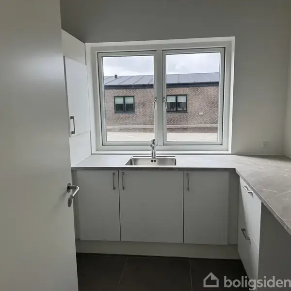 En køkkenvask med hvidt skab under er centralt placeret foran et vindue. Omgivet af beige bordplader og set mod en murstensbygning udenfor vinduet.
