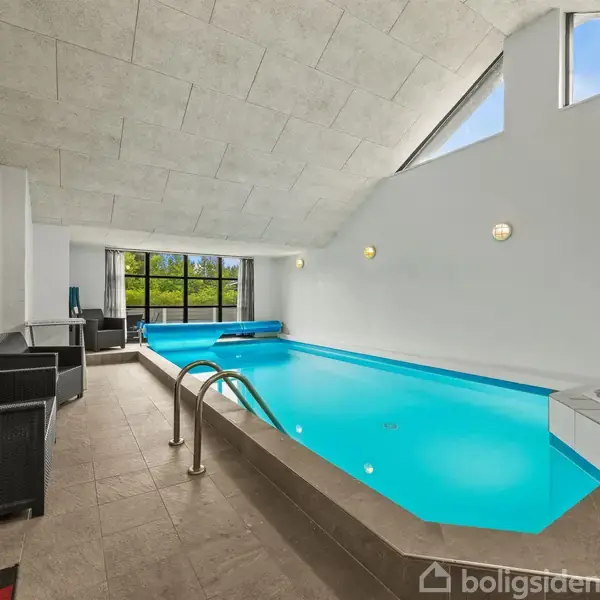 Et indendørs swimmingpoolområde med en lys blå pool, omgivet af fliser. Ved siden af poolen er der en rund jacuzzi. Rummet har et skråt loft og store vinduer med udsigt til træer.