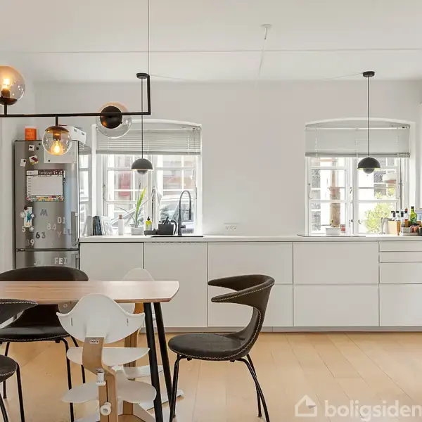 Et moderne køkken med hvide skabe og indbyggede ovne i en lys lejlighed. En spisebordsgruppe med forskellige stole står midt i rummet. Store vinduer giver naturligt lys. En køleskab med magneter ses i baggrunden.