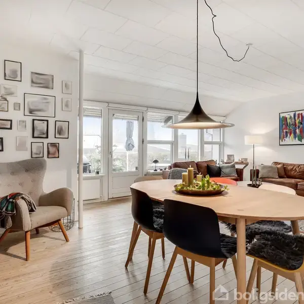 Et lyst stueområde med en rund spisebord omgivet af fire stole. Baggrunden indeholder en væg med indrammet kunst og billeder, en lænestol, og en stor brun sofa. En hængelampe hænger over bordet.