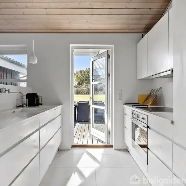 Et hvidt, moderne køkken med indbyggede skabe i minimalistisk design åbner op mod en terrasse gennem en glasdør, hvor der er udsigt til en grøn have.