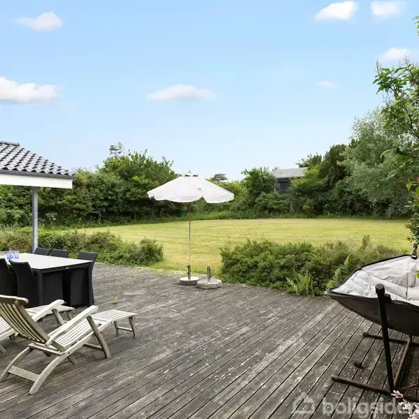 Terrasse med liggestole og spisebord står under en parasol. Omgivet af en velholdt græsplæne og grønne buske, ligger ved siden af et sortmalet hus med skifertag.