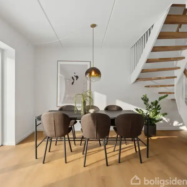 En moderne spisestue med et bord og seks brune stole står ved siden af en stor glasdør. Over bordet hænger en gylden lampe. En trappe leder til en anden etage.
