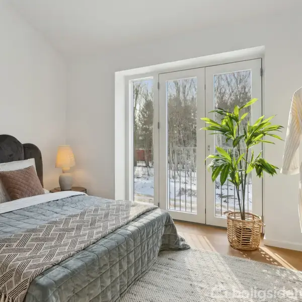 Soveværelse med en dobbeltseng dækket af et gråt sengetæppe, placeret ved siden af en lampe. Glasdøre fører til en sneklædt have. En plante og tøjstativ står i nærheden.