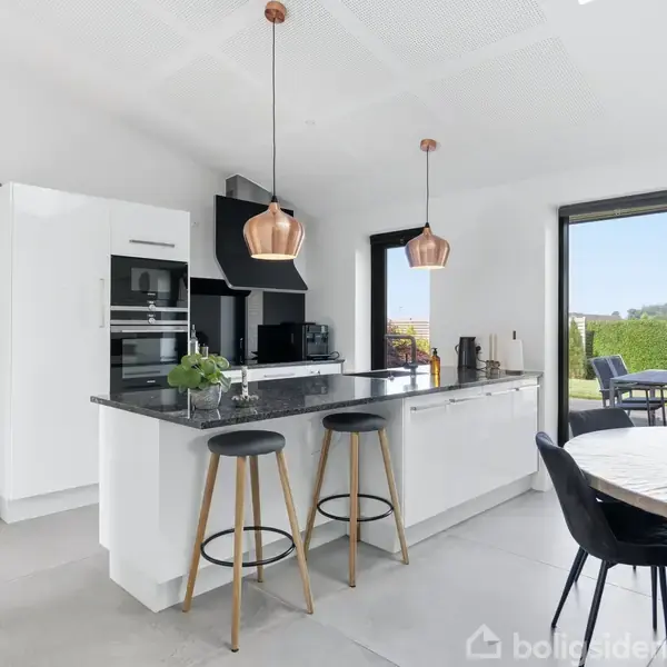 Et moderne køkken med en stor ø med to barstole. Hvide skabe og moderne apparater ses i baggrunden. To kobberfarvede lamper hænger over øen. Glidende dør fører til terrasseområde.