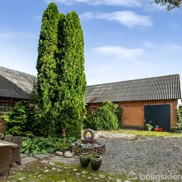 Sammenstillingen af et gammelt murstenshus og en lade, med et højt cyprestræ imellem dem, skaber et idyllisk gårdhavemiljø. Møbler i kurveflet står på terrassen.