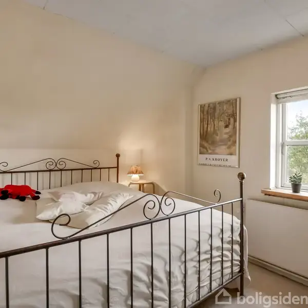 En metalseng står i et lyst soveværelse med natlamper og et billede på væggen. To små planter står i vindueskarmen ved siden af et stort vindue.