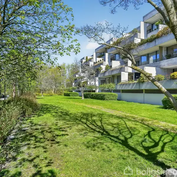 Beboelsesejendom med altaner, stille beliggende i et grønt område omkranset af træer og buske. Solrigt vejr med skarpe skygger på græsset fra træerne. Ingen tekst bemærket.
