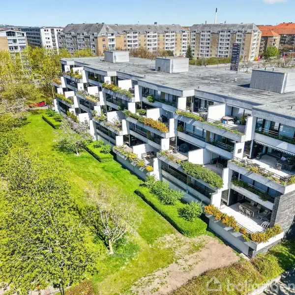 En modern lejlighedsbygning med flere etager har balkoner fyldt med planter. Den står i et rekreativt område omgivet af grønne træer og græs med andre boligbygninger i baggrunden.