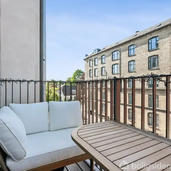Kaffebord og sofa med hvide hynder stående på en balkon. Omgivet af potteplante og trægelænder, set mod en baggrund af en murstensbygning og trætoppe under en klar blå himmel.