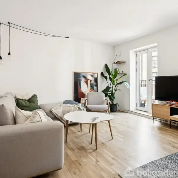 En lys stue med gråt hjørnesofa placeret til venstre, et rundt hvidt sofabord i midten, og et fladskærms-TV til højre på et lavt skab. En grøn plante står i hjørnet ved altandøren.