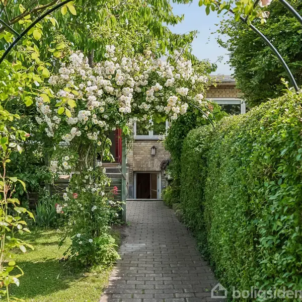 En murstensbygning med et indgangsparti ses i baggrunden. En blomstrende bue af hvide blomster strækker sig over en flisebelagt sti omgivet af grønne hække og velplejede planter.