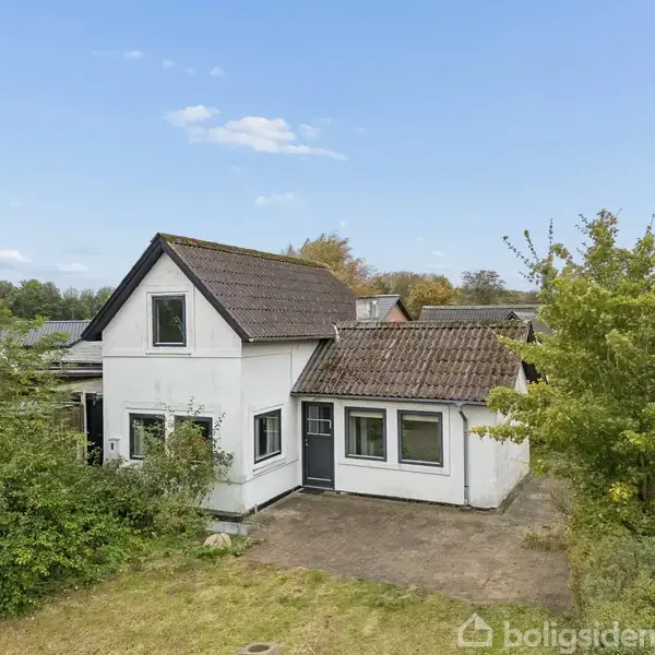 Et hvidt hus med brun tag udført til boligformål står stille i en have med grønne buske og træer. Huset har en dør foran og ligger i et roligt nabolag.