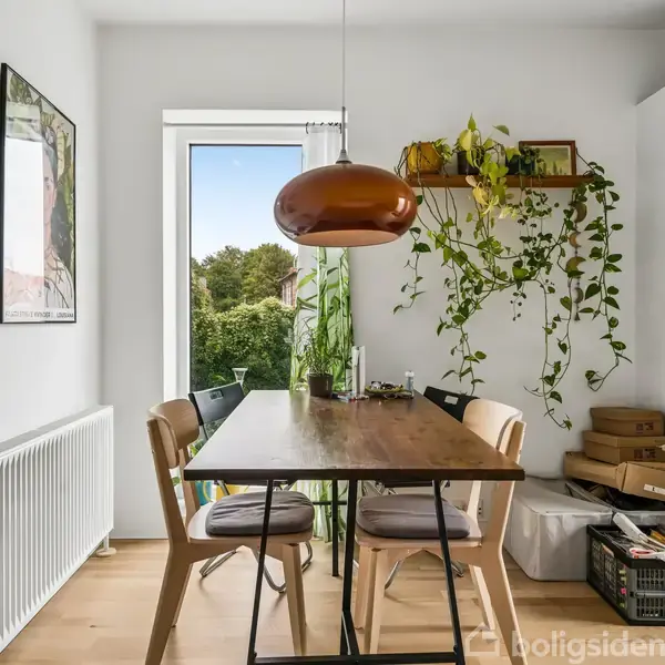 Et spisebord med fire stole står under en brun pendellampe; omgivet af vægkunst, planter og en stor hvid skab ved siden af et vindue, der viser en grøn have.