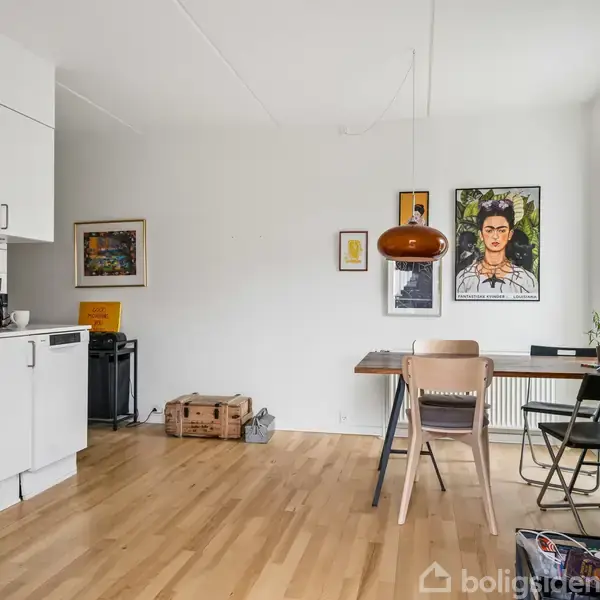 Køkkenområde med hvide skabe og en opvaskemaskine til venstre. Foran står et spisebord med to stole under en kobberfarvet loftlampe. Væggene har kunstværker og plakater.