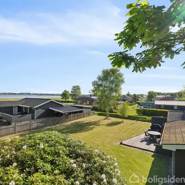 Et sommerhus med en sort facade står stille i en grøn have. Sollys oplyser området, omgivet af andre huse og en fjord i baggrunden. Træer giver skygge.