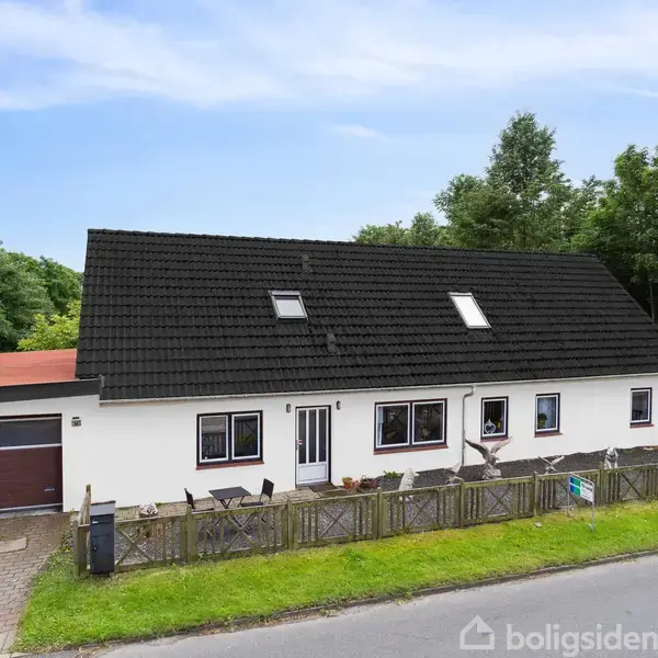 Et hvidt enfamiliehus med sort tag og flere vinduer står ved en vej. Huset har en garage til venstre, omkranset af en grøn have og træer i baggrunden.