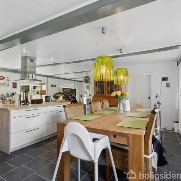 Et moderne køkken med en stor træbord, omgivet af hvide stole. Grønne lamper hænger over bordet. Køkkenøen har hvide skabe, og rummet har sorte flisegulve og hvide vægge.