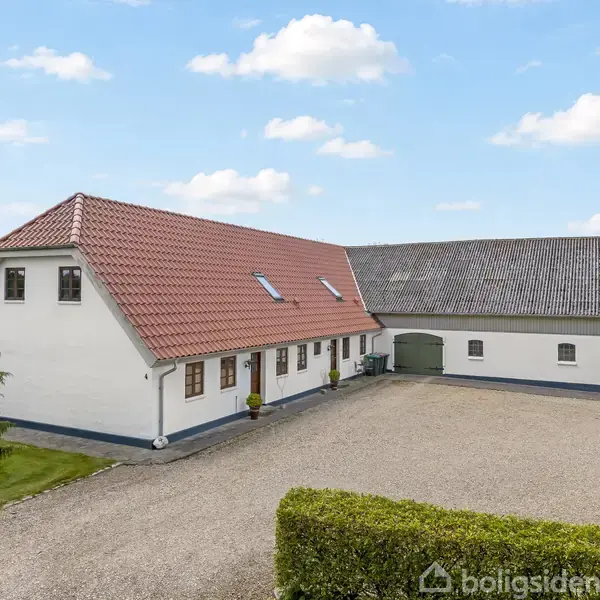 Et hvidt landhus med rødt tegltag og flere små vinduer står på en grusbelagt gårdsplads, omgivet af grønne marker og hække under en blå himmel med skyer.