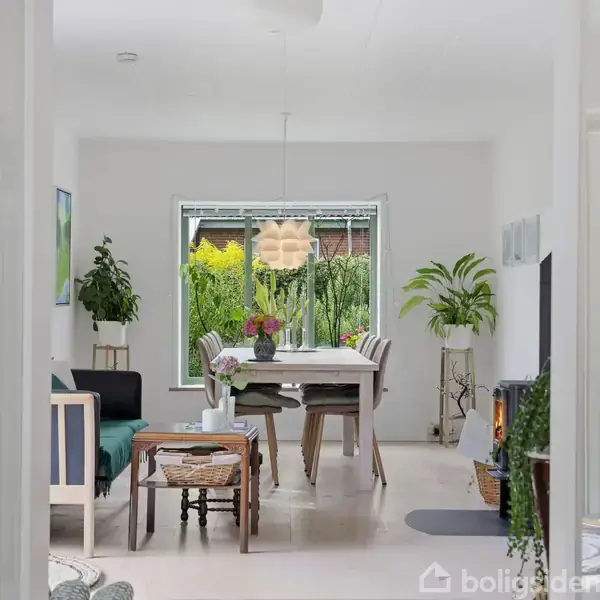 Stue med spisebord og stole, omgivet af planter og dekorationer. Et stort vindue viser en grøn have udenfor. Rummet er lyst og indbydende med moderne indretning.