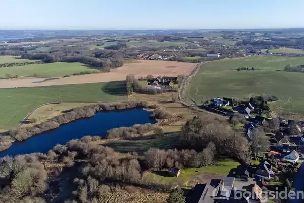 En sø omgivet af træer ligger stille i en landlig by, hvor små huse og marker strækker sig i det fjerne under en klar blå himmel.