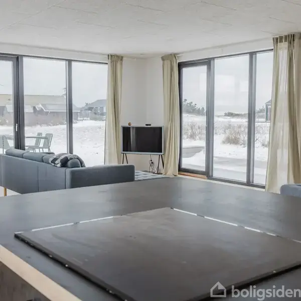 Stue med mørkt bord i forgrunden, sofa og tv i midten, omgivet af store vinduer med udsigt til sneklædt landskab udenfor.