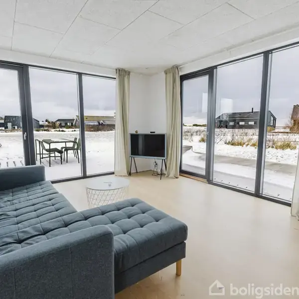 Blå sofa står i en lys stue med store vinduer. Udenfor ses et snedækket landskab med nogle huse. Et lille fjernsyn og et bord findes nær vinduerne.