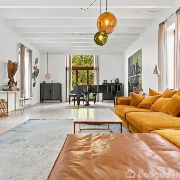 Stor stue med gul sofa arrangeret i et hjørne, moderne kunst på væggene, pendellamper og et flygel i baggrunden nær store vinduer og gardiner, der slipper naturligt lys ind.