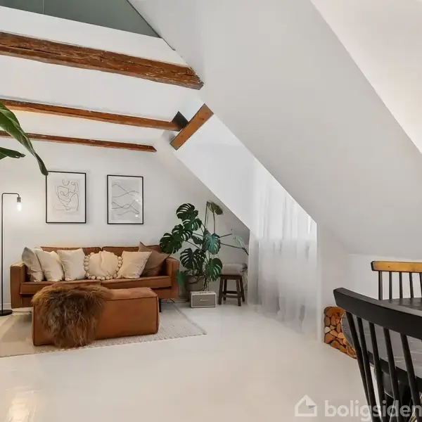 En brun lædersofa står på et tæppe, omgivet af planter og kunst på væggen under skrå loftsbjælker. En gulvlampe lyser op i rummet med lyse, moderne møbler.