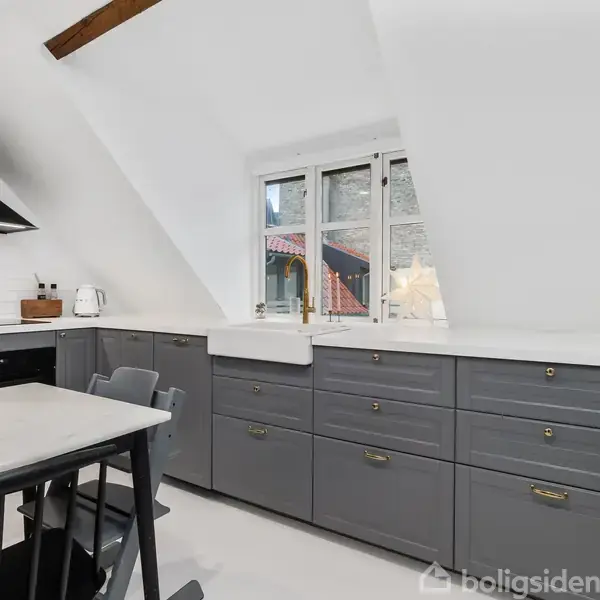 Et lyst køkken med grå skabe og hvid bordplade. Bord med sorte stole står ved siden af. Vindue med udsigt til en mur omslutter rummet, og der er træbjælker i loftet.