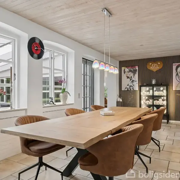 Brun hjørnesofa står langs en stor stueopdeling med gyldne lameller; sofabord foran. Store vinduer slipper lys ind, og en stor plante står i hjørnet på gråt klinkegulv.