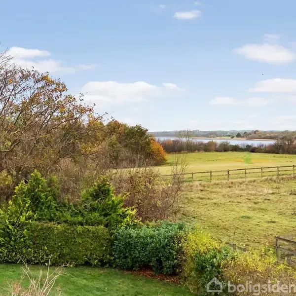 Grønne buske og træer står stille i forgrunden, mens en mark strækker sig mod en sø i baggrunden, omkranset af en lav hegn i et åbent landskab.