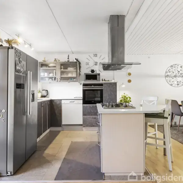 Stue med en grå sofa og moderne gyngestol, vender mod stor fladskærm. Spisebord med otte stole i baggrunden, tilstødende køkken ses. Indretning er hyggelig med lys træfarve.