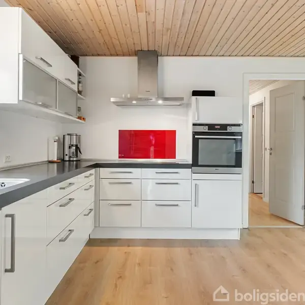 Et moderne køkken med hvide skabe og sort bordplade. En indbygget ovn og emhætte findes ved en rød backsplash. Trægulv og træloft udgør rummet.