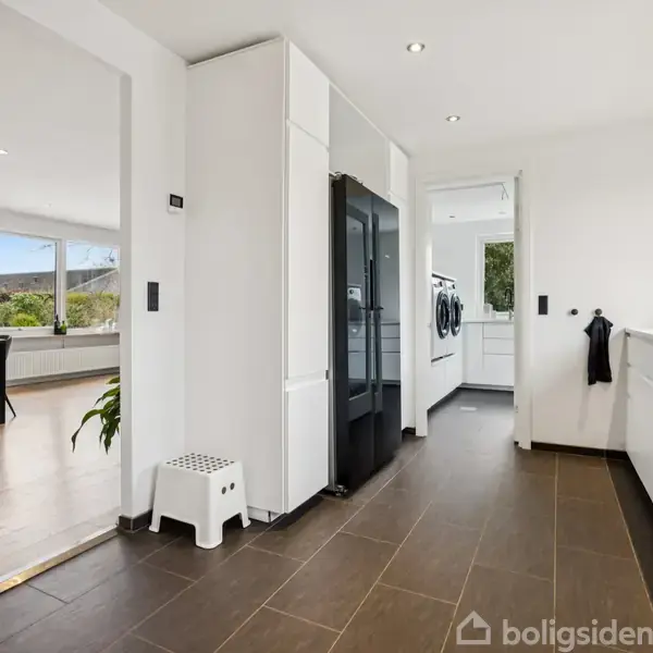 Køkken med hvide skabe og en sort køleskab. En sort kran står på bordpladen. I baggrunden ses et spisebord og trægulv. En lille skammel står ved et skab.