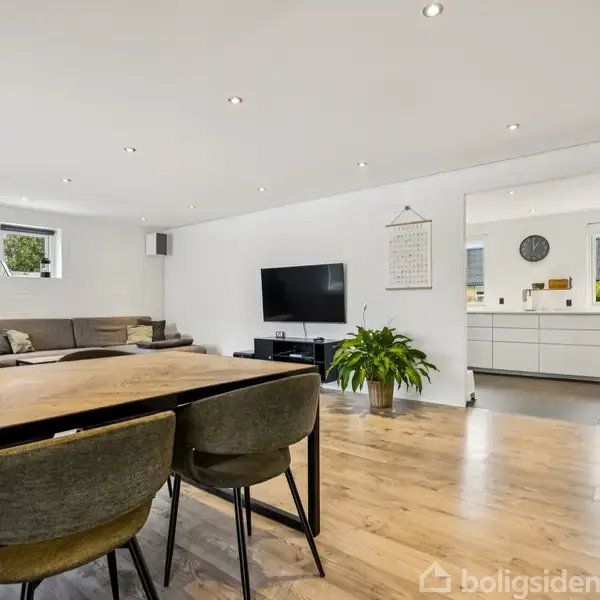 Stor stue med spisebord og stole foran. Sofa og fjernsyn på en væg. I baggrunden ses et moderne køkken med hvide skabe og vinduer. Indendørs plante ved fjernsynet.