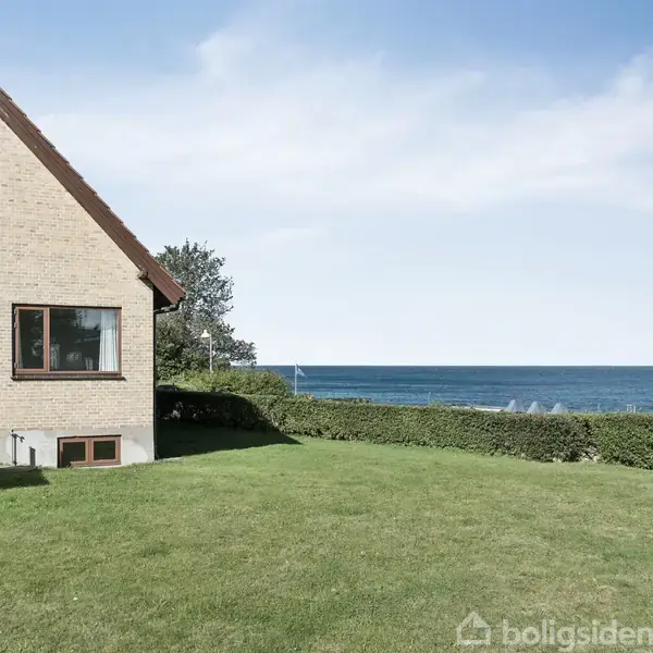 Hus med rødt tag står stille ved siden af en velplejet plæne, omgivet af en hæk. I baggrunden ses havet under en klar himmel.