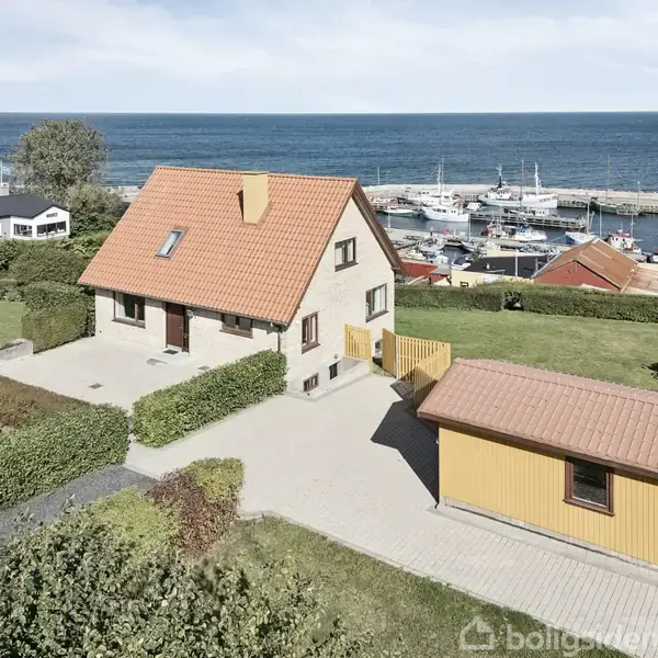 Et rødt hus med gavl og rødt tegltag står i en velplejet have. Ved siden af ses en gul bygning. I baggrunden ligger en havn med både og havudsigt.