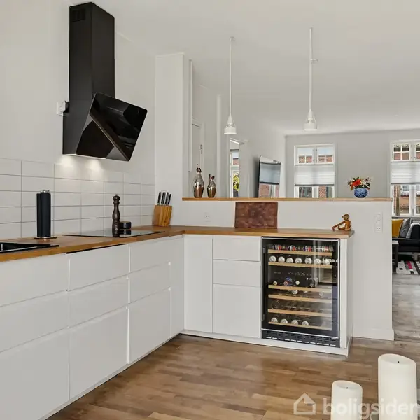 Et moderne køkken med hvide skabe og en træbordplade. En vinreol er indbygget under disken. I baggrunden er der spise- og opholdsområde med vinduer.