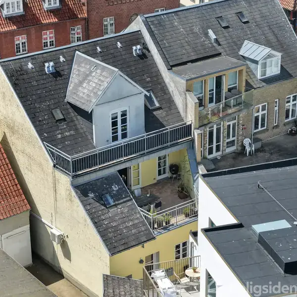 Et fleretagers byhus har en stor balkon med møbler på øverste etage. Omkringliggende bygninger inkluderer røde murstenshuse og moderne strukturer. Stilen er blandet mellem traditionel og moderne arkitektur.