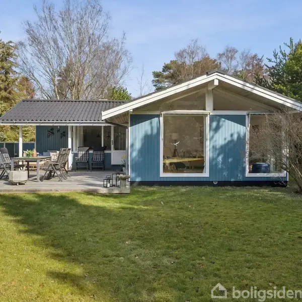 Blå træsommerhus med store vinduer står i en grøn have. Terrasse foran huset har havemøbler. Omgivet af træer og buske, skaber en rolig, naturlig atmosfære.